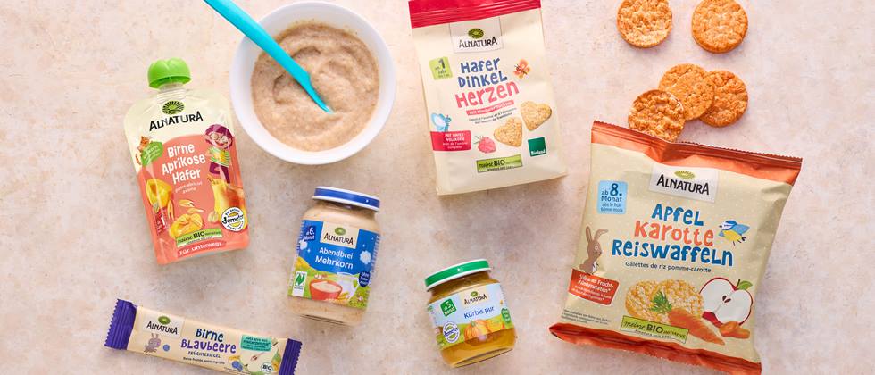 Alnatura Babykostprodukte, darunter Babybrei, Fruchtmus, Kekse und Reiswaffeln, liegen auf einem beigefarbenen Hintergrund, daneben einige blaue Beeren.