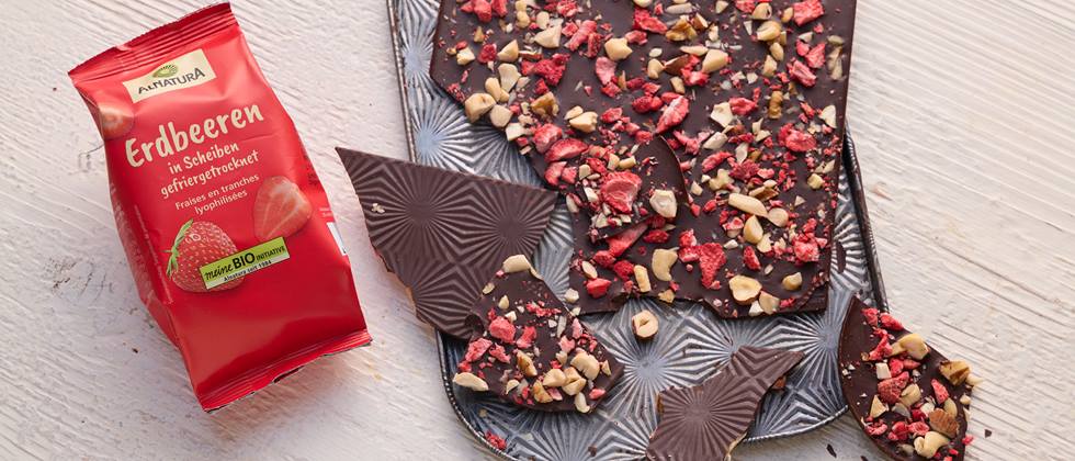 Auf einem hellen Holz-Untergrund liegt eine Packung Alnatura gefriergetrocknete Erdbeeren, daneben ist ein kleines Backblech. Auf dem Backblech liegt Schokolade bestreut mit gefriergetrockneten Erdbeeren und gehackten Nüssen. Einige Stücke sind von der Schokolade abgebrochen und liegen ebenfalls auf dem Blech.