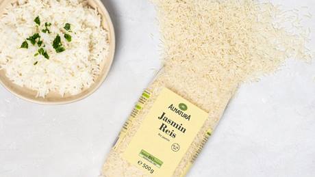 Eine Packung Alnatura Jasmin Reis liegt aufgeschnitten und der Reis liegt verteilt auf grauem Untergrund, daneben steht ein Teller gekochter Reis