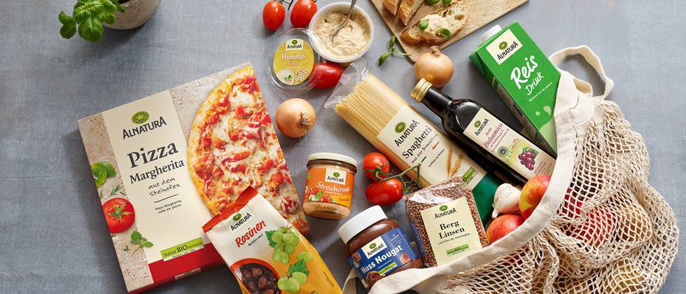 Eine Auswahl von Alnatura-Produkten liegen auf einem grauen Untergrund. Darunter Pizza Margherita, Rosinen, Streichcreme Toskana, Hummus, Spaghetti, Balsamico, Reis Drink, Nuss Nougat Creme, Berglinsen, Brot, Tomaten, Zwiebeln, Knoblauch und Äpfel. Neben der Pizza steht ein Topf mit frischem Basilikum. Die Produkte sind teilweise in einer Jutetasche drapiert.
