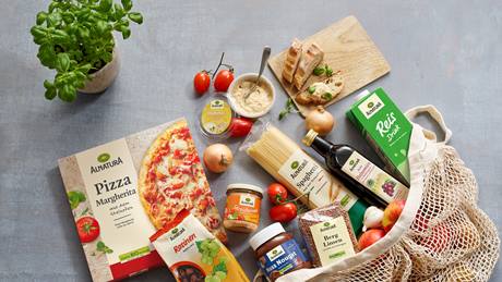 Eine Auswahl von Alnatura-Produkten liegen auf einem grauen Untergrund. Darunter Pizza Margherita, Rosinen, Streichcreme Toskana, Hummus, Spaghetti, Balsamico, Reis Drink, Nuss Nougat Creme, Berglinsen, Brot, Tomaten, Zwiebeln, Knoblauch und Äpfel. Neben der Pizza steht ein Topf mit frischem Basilikum. Die Produkte sind teilweise in einer Jutetasche drapiert.