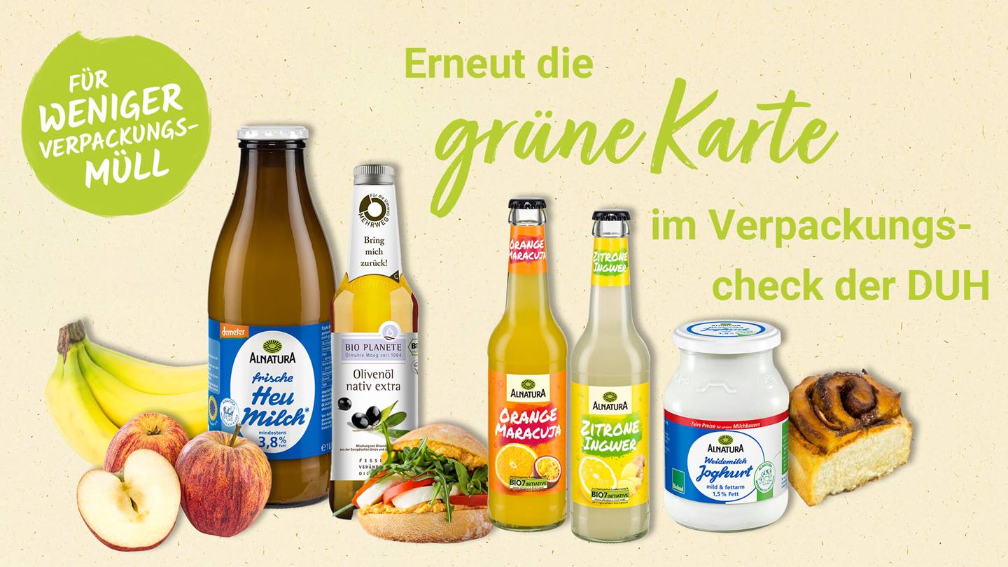 Eine Produktpräsentation mit verschiedenen Produkte von Alnatura, darunter Bananen, Äpfel, ein belegtes Brötchen, eine halbe Zimtschnecke, Heumilch, Orangensaft-Maracuja-Saft, Zitronen-Ingwer-Saft, Joghurt und ein Olivenöl von Bio Planete stehen vor einem beigen Hintergrund. Es wird für weniger Verpackungsmüll geworben und die "Grüne Karte" im Verpackungcheck der DUH hervorgehoben. Das Gesamtbild vermittelt einen Eindruck von Nachhaltigkeit.