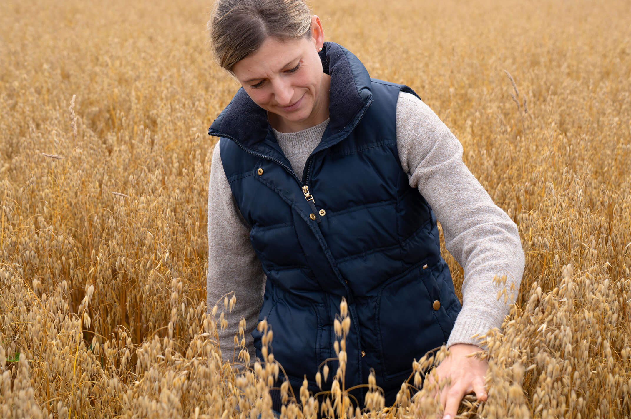 Eine Frau mit blauer Daunenweste und grauem Strickpullover steht in einem Haferfeld und streicht über die Ähren.