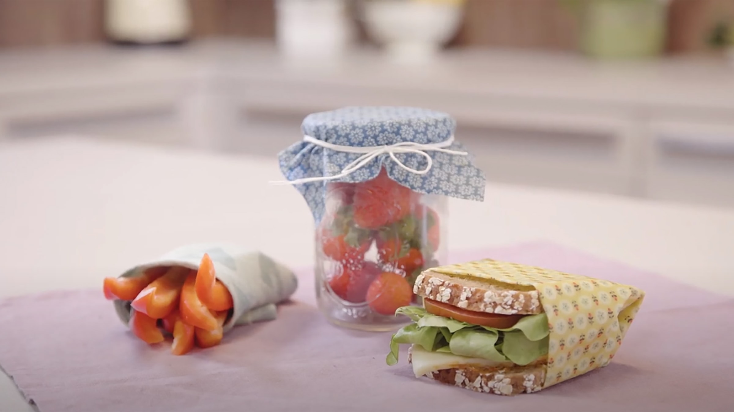 Video Startbild: Es sind drei verschiedene Beispiele für die Verpackung von Lebensmitteln mit Wachstüchern zu sehen. Karotten und ein belegtes Brot in jeweils einem Wachstuch eingewickelt. Sowie ein Glas, dass mit einem Wachstuch verschlossen wurde.