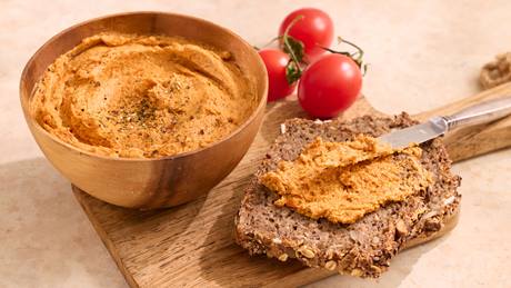 Holzschale mit Tomatenbutter neben Vollkornbrot mit Tomatenbutter bestrichen und zwei Tomaten im Hintergrund.