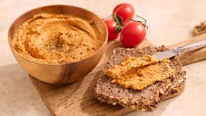 Holzschale mit Tomatenbutter neben Vollkornbrot mit Tomatenbutter bestrichen und zwei Tomaten im Hintergrund.