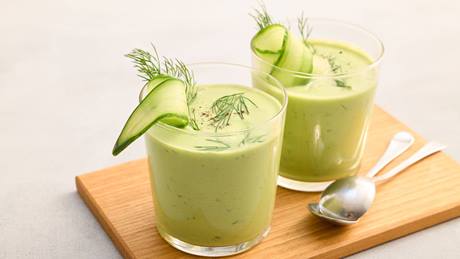 Zwei Gläser mit grüner kalter Gurkensuppe, garniert mit Gurkenscheiben und Dill, auf einem Holzbrett mit zwei Löffeln.