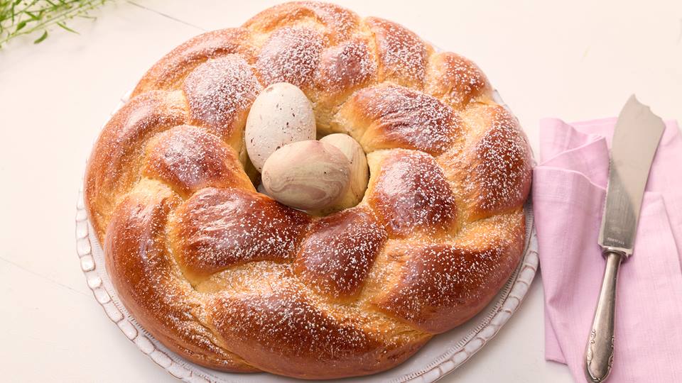 Auf einer runden Kuchenplatte aus Porzellan liegt ein runder Hefezopf bestreut mit Puderzucker. In der Mitte des Hefezopfes liegen marmorierte Ostereier. Neben dem Hefezopf liegt ein Kuchenmesser und eine rosafarbene Stoffserviette.