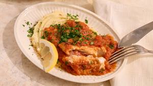 Teller mit Fischfilets in Tomatensauce auf Kartoffelpüree garniert mit gehackter Petersilie und Zitronenscheibe, daneben Messer und Gabel.