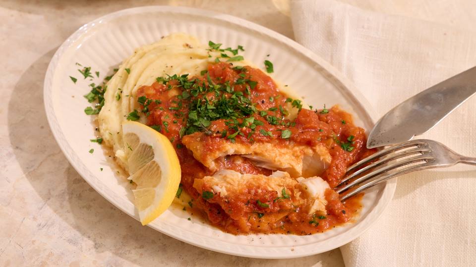 Teller mit Fischfilets in Tomatensauce auf Kartoffelpüree garniert mit gehackter Petersilie und Zitronenscheibe, daneben Messer und Gabel.