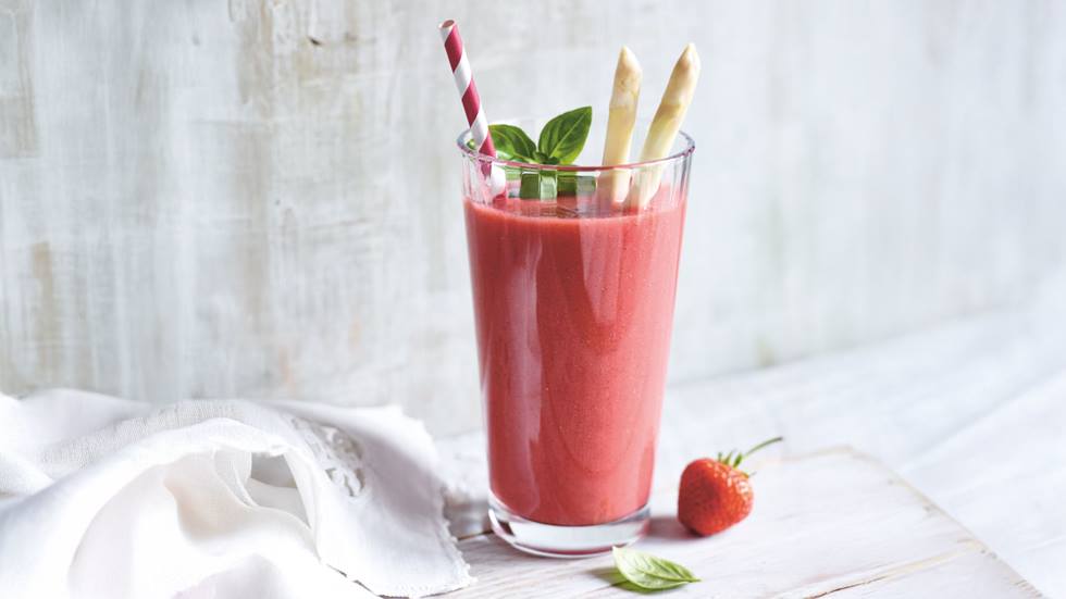 Ein Glas mit einem rosa Erdbeer-Spargel-Smoothie mit Vanille. Der Smoothie ist mit zwei Stangen weißen Spargels und einem Basilikumblatt garniert. Ein roter Strohhalm steckt im Getränk. Im Vordergrund liegt eine einzelne Erdbeere. Im Hintergrund ist eine Stoffserviette zu sehen. Das Ganze wird auf einem hellen Holztisch präsentiert. Es wirkt frisch, appetitlich und sommerlich.