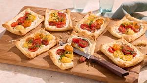 Mehrere rechteckige Tartelettes mit Blätterteig, Frischkäse und halbierten roten und gelben Kirschtomaten auf einem Holzbrett, daneben ein Messer mit braunem Griff.