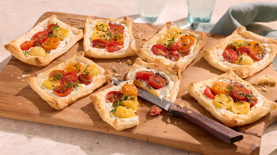 Mehrere rechteckige Tartelettes mit Blätterteig, Frischkäse und halbierten roten und gelben Kirschtomaten auf einem Holzbrett, daneben ein Messer mit braunem Griff.