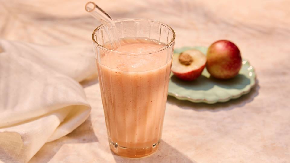 Glas mit cremigem Nektarinensmoothie und Glasstrohhalm neben Teller mit halbierter und ganzer Nektarine.