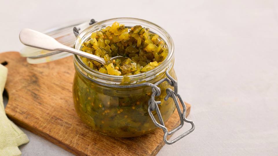 Offenes Glas mit gewürfeltem Gurkenrelish und einem Löffel auf einem Holzbrett.