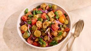 Schüssel mit Tomaten-Brotsalat aus halbierten roten und gelben Kirschtomaten, Brotwürfeln, roten Zwiebelringen und grünen Kräutern, daneben goldfarbener Löffel und Gabel.