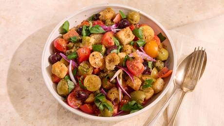 Schüssel mit Tomaten-Brotsalat aus halbierten roten und gelben Kirschtomaten, Brotwürfeln, roten Zwiebelringen und grünen Kräutern, daneben goldfarbener Löffel und Gabel.