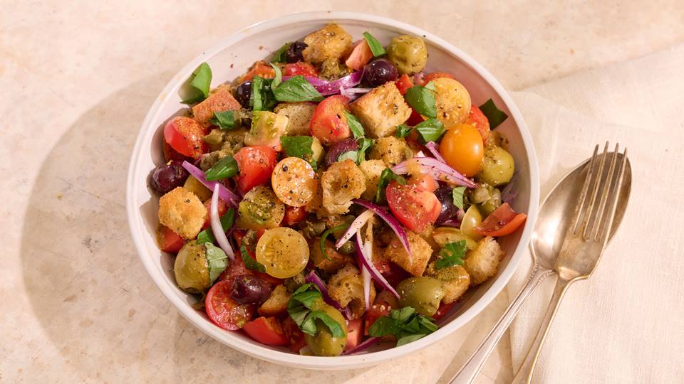 Schüssel mit Tomaten-Brotsalat aus halbierten roten und gelben Kirschtomaten, Brotwürfeln, roten Zwiebelringen und grünen Kräutern, daneben goldfarbener Löffel und Gabel.