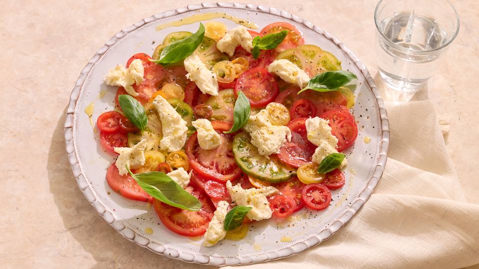 Teller mit Tomatenscheiben, Mozzarellastücken und Basilikumblättern, daneben ein Glas Wasser auf einem hellen Tuch.