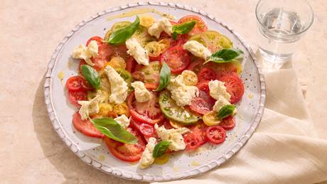 Teller mit Tomatenscheiben, Mozzarellastücken und Basilikumblättern, daneben ein Glas Wasser auf einem hellen Tuch.