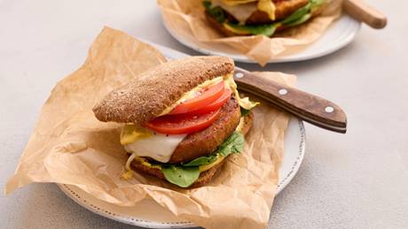 Burger mit Vollkornbrötchen, Linsen-Patty, Tomatenscheiben, Salat und Sauce auf einem weißen Teller mit Backpapier und Messer.