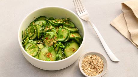Schüssel mit Gurkenscheiben, Sesam und Frühlingszwiebeln, daneben kleine Schale mit Sesam und eine Gabel auf hellem Untergrund.