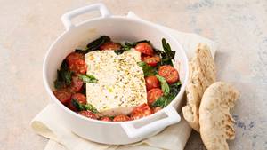 Ofenfeta in einer weißen Auflaufform mit Kirschtomaten, Spinat und Gewürzen daneben zwei Fladenbrote.