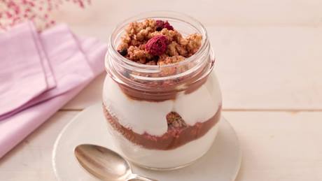 Auf einem kleinen weißen Porzellantellerchen steht ein bauchiges Schaubglas, daneben liegt ein silberner Teelöffel. In dem Glas ist ein Dessert geschichtet. Unten im Glas eine Schicht Joghurt, darüber rotes Fruchtpüree mit Frucht-Crunchy, erneut eine Schicht Joghurt und als oberste Schicht wieder Frucht-Crunchy mit rotem Fruchtpüree. Eine getrocknete Himbeere und Johannisbere liegen oben.