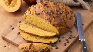 Auf einem Holzbrett liegt ein aufgeschnittenes Kürbisbrot mit Kernen. Weitere Kürbiskerne sind verstreut, neben dem Brot liegt ein Messer, ein aufgeschnittener halber Hokkaido-Kürbis und eine braune Stoffserviette.