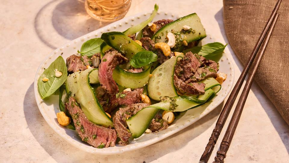 Teller mit thailändisch gewürztem Rindfleischsalat aus dünnen Rindfleischstreifen, Gurkenscheiben, Cashewnüssen und Basilikumblättern, daneben Essstäbchen und ein Glas Getränk