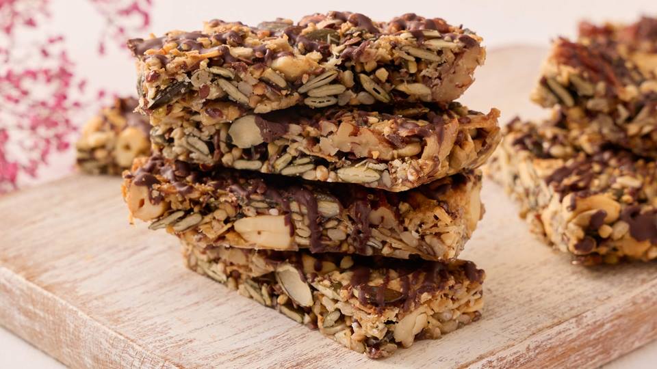 Ein Stapel von vier Schoko-Müsliriegeln ist auf einem hölzernen Schneidebrett. Hinter dem Stapel liegen noch weitere Schoko-Müsliriegel.