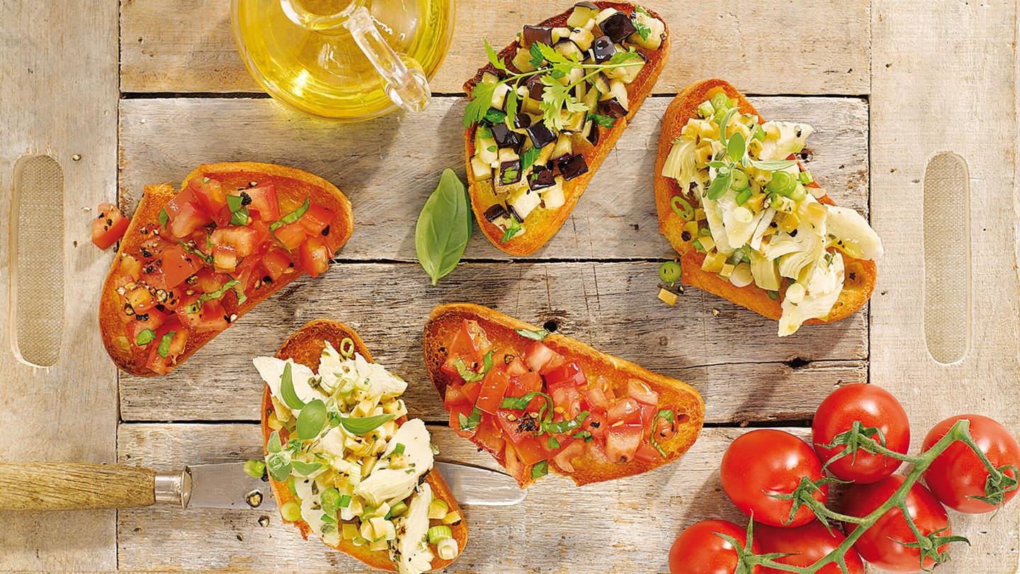 Auf einem Holzbrett liegen fünf Schreiben Bruschetta in drei verschiedenen Varianten Bruschetta. Es ist eine kleine Kanne Öl und Rispentomaten zu sehen.