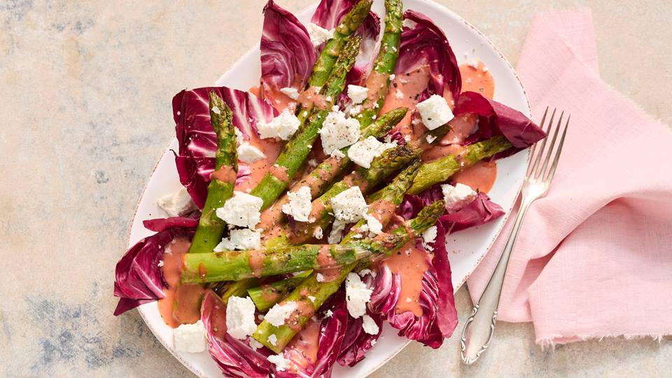 Grüner Spargel auf Radicchio mit Fetawürfeln und rosa Sauce auf weißem Teller neben silberner Gabel und rosa Stoffserviette.