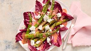 Grüner Spargel auf Radicchio mit Fetawürfeln und rosa Sauce auf weißem Teller neben silberner Gabel und rosa Stoffserviette.