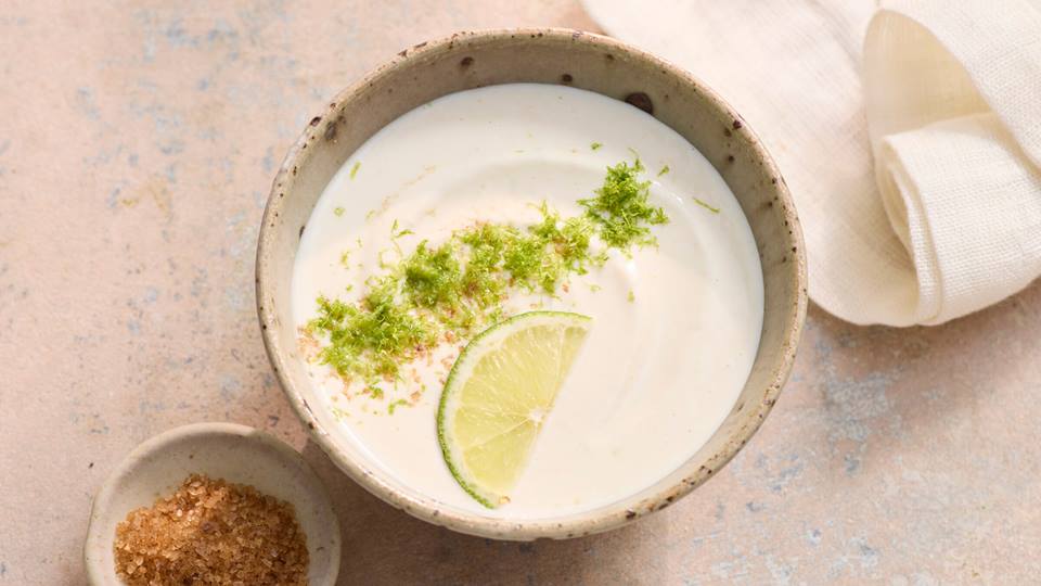 Schale mit weißem Joghurt-Limetten-Dip, garniert mit Limettenscheibe und Limettenabrieb, daneben kleine Schale mit braunem Gewürz und weißes Tuch.