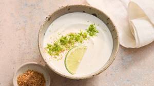 Schale mit weißem Joghurt-Limetten-Dip, garniert mit Limettenscheibe und Limettenabrieb, daneben kleine Schale mit braunem Gewürz und weißes Tuch.