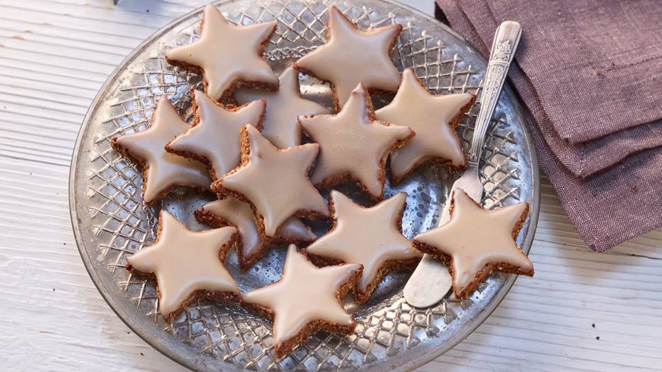 Viele gebackene Zimtsterne liegen auf einem verzierten, silbernen Teller, dazu ein silbernes Buttermesser. Die Kekse sind mit einer Zucker-Glasur überzogen. Neben dem Teller liegt eine braune Stoffserviette. Der Hintergrund besteht aus einer weißen, holzähnlichen Oberfläche.