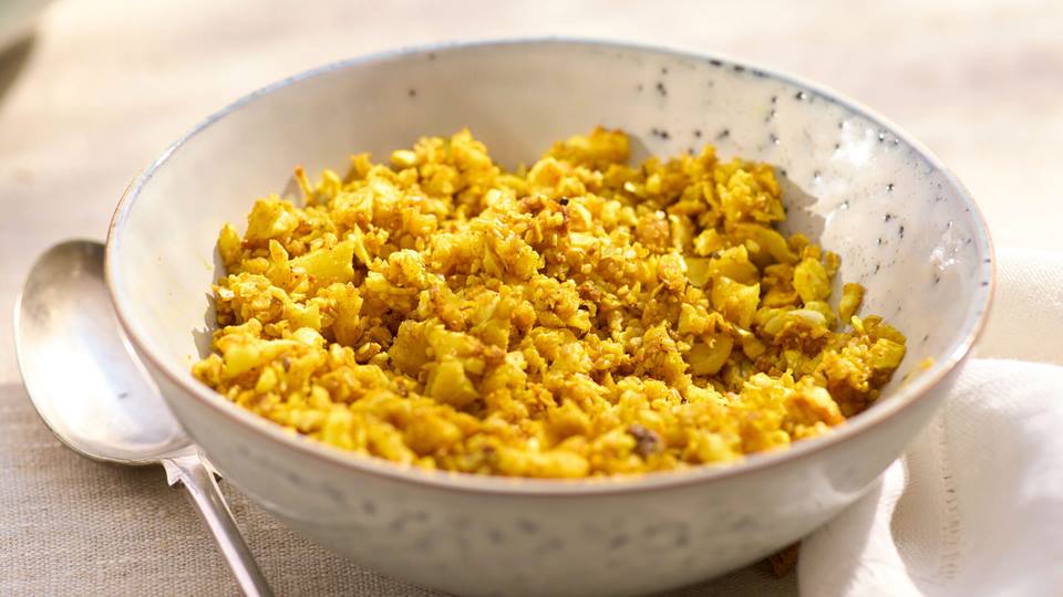 Schale mit Blumenkohl und Sesambröseln, daneben ein Löffel. Auf der anderen Seite eine weiße Serviette, im Hintergrund unscharf eine große Schüssel mit Salat.