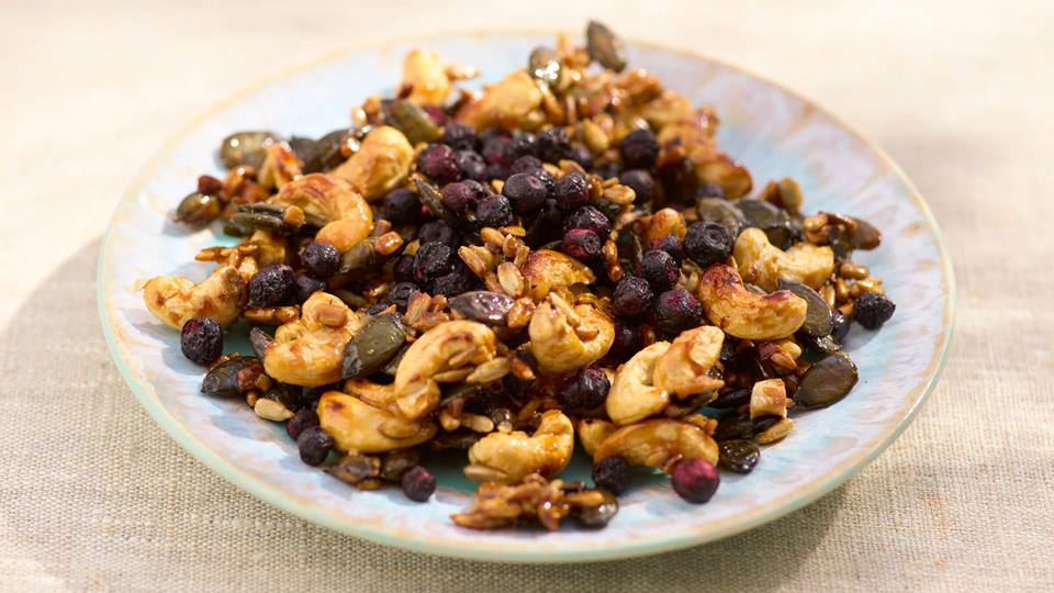 Teller mit Honig-Salz-Kernen, dazu Kürbiskerne und getrockneten Beeren auf einem hellen Stoffuntergrund.
