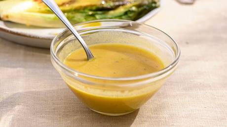 Glas mit Honig-Senf-Dressing​ und Löffel davor auf hellem Tischtuch, im Hintergrund Teller mit gegrilltem Gemüse und daneben Besteck.