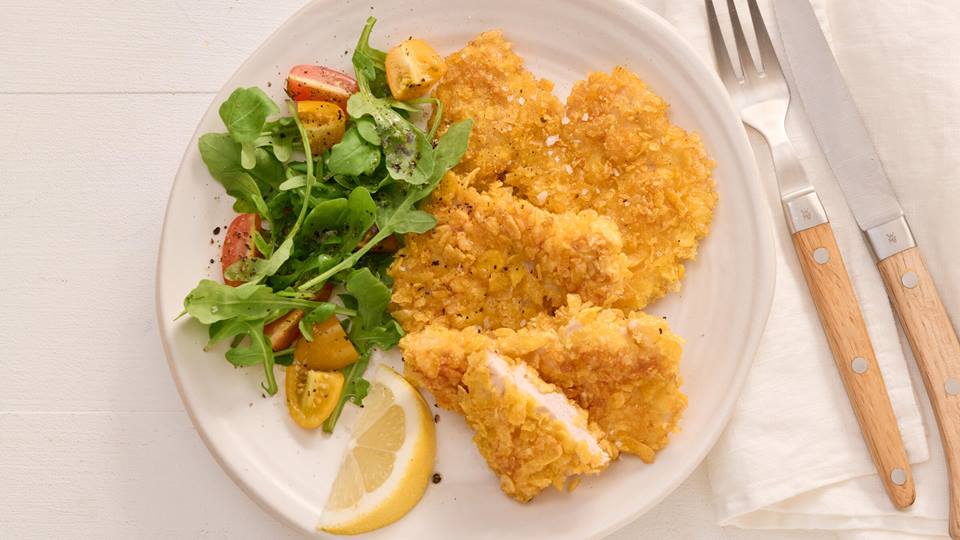 Auf einem weißen Porzelleanteller liegt ein Salatbouquet, ein Zitronenschnitz und vier Mini-Schnitzel mit Cornflakes-Kruste. Ein Mini-Schnitzel ist aufgeschnitten. Neben dem Teller liegt eine Gabel, ein Messer und eine weiße Stoffserviette.