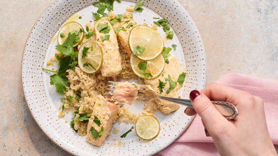Hand mit Gabel nimmt ein Stück gebackenen Lachs von einem Teller mit Couscous, Limettenscheiben und Koriander.