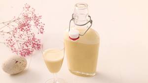 Auf einem weißen Holztisch steht eine kleine Glasflasche mit Bügelverschluss und ein Glas mit Eierlikör. Daneben liegt ein Osterei und rosa Schleierkraut.