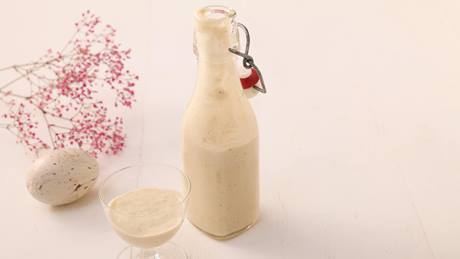 Auf einem weißen Holztisch steht eine kleine Glasflasche mit Bügelverschluss und ein Glas mit veganem Eierlikör. Daneben liegt ein Osterei und rosa Schleierkraut.