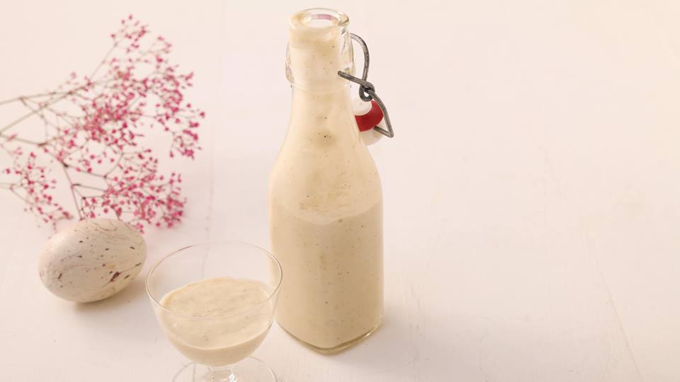 Auf einem weißen Holztisch steht eine kleine Glasflasche mit Bügelverschluss und ein Glas mit veganem Eierlikör. Daneben liegt ein Osterei und rosa Schleierkraut.