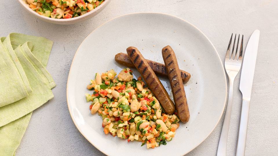 Drei gegrillte Würstchen neben einem Bohnensalat mit gehacktem Gemüse auf weißem Teller mit Besteck und grünem Stoffserviette.