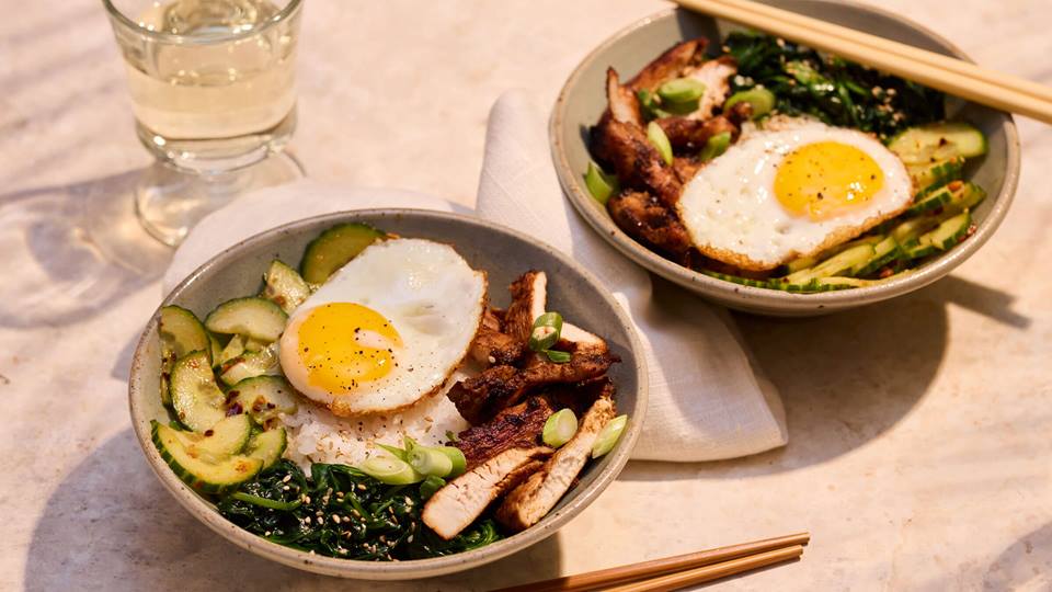 Zwei Schalen mit Bibimbap, jeweils mit Spiegelei, Gurkenscheiben, gebratenem Fleisch und grünem Gemüse, daneben Essstäbchen und ein Glas Wasser.