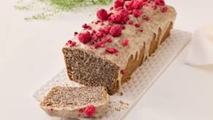 Auf hellem Untergrund steht eine weiße Kuchenplatte aus Porzellan. Auf der Platte ist ein Kastenkuchen. Der Kuchen ist angeschnitten und das erste Stück liegt noch auf der Kuchenplatte. Das innere des Kuchens ist zu sehen. Im Kuchen ist Blaumohn verbacken. Der Kuchen hat eine helle Glasur und ist mit leuchtend roten gefriergetrockneten Himbeeren bestreut.