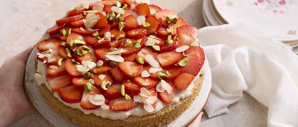 Erdbeerkuchen mit Sahne, Mandelsplittern und Pistazienstückchen auf einem Teller, gehalten von zwei Händen.
