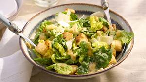 Schüssel Ceasar Salat mit Harissa Dressing​ und zwei Gabeln auf hellem Tisch mit Glasflaschen und Zitronen im Hintergrund.